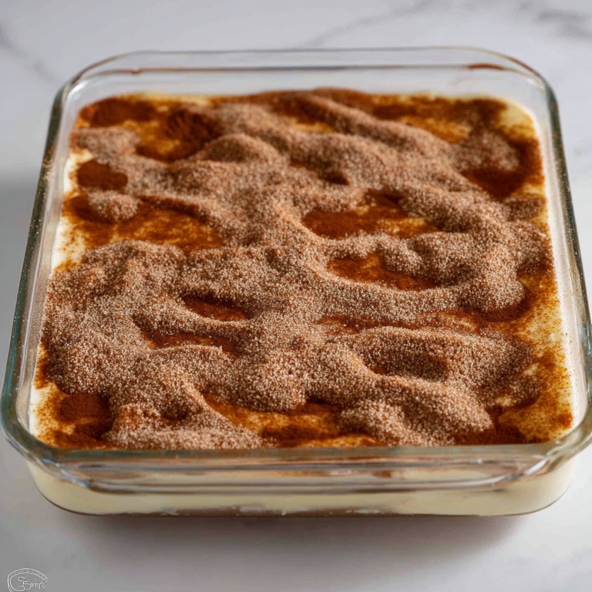 Churro cheesecake in a glass baking dish topped with a cinnamon-sugar layer, ready to bake into a sweet, layered dessert.