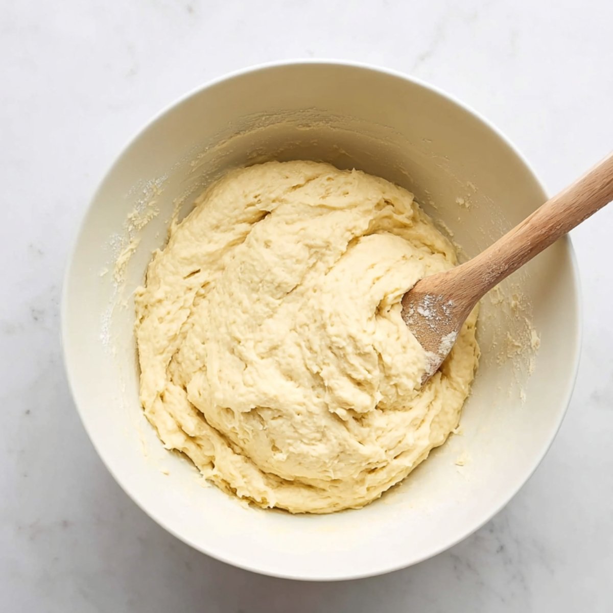 Overhead view of a bowl with naan dough being mixed with a wooden spoon. The dough is thick and sticky, with a smooth, slightly elastic texture, ready for kneading and resting.