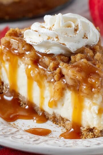 A slice of apple crisp cheesecake topped with whipped cream, caramel sauce, and cinnamon, showing a creamy interior with a graham cracker crust.