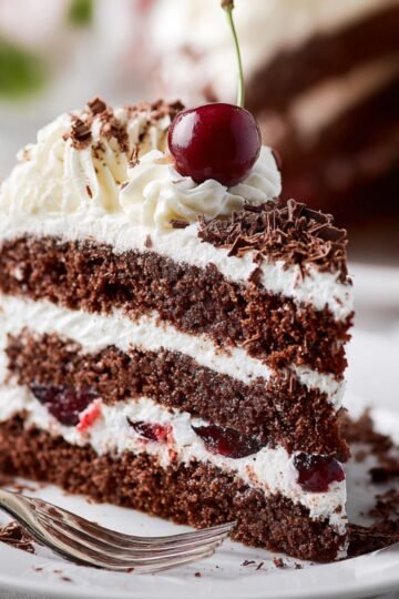 Slice of Black Forest cake with chocolate layers, whipped cream, cherries, and chocolate shavings on a plate.