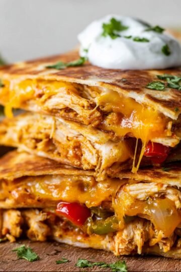 Stack of Chicken Quesadilla filled with shredded chicken, melted cheese, and bell peppers, topped with a dollop of sour cream and fresh cilantro, served with guacamole on the side.