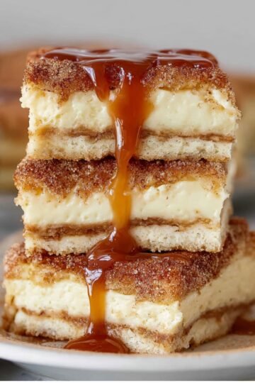 Sliced churro cheesecake bars stacked on a plate, with caramel sauce dripping down the sides, showcasing their creamy cheesecake filling and crispy cinnamon-sugar crust.