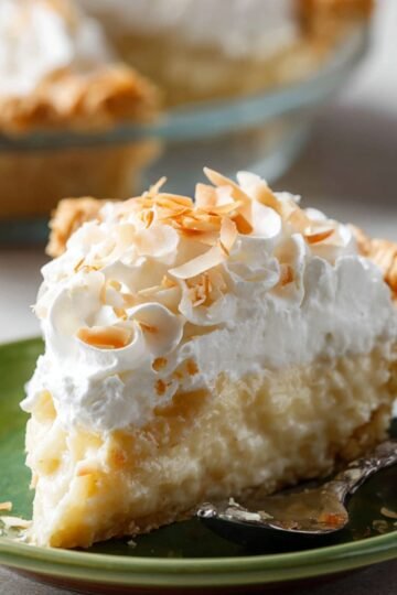 Slice of coconut cream pie on a green plate with whipped cream, toasted coconut flakes on top, and a silver spoon beside it. The whole pie is visible in the background.