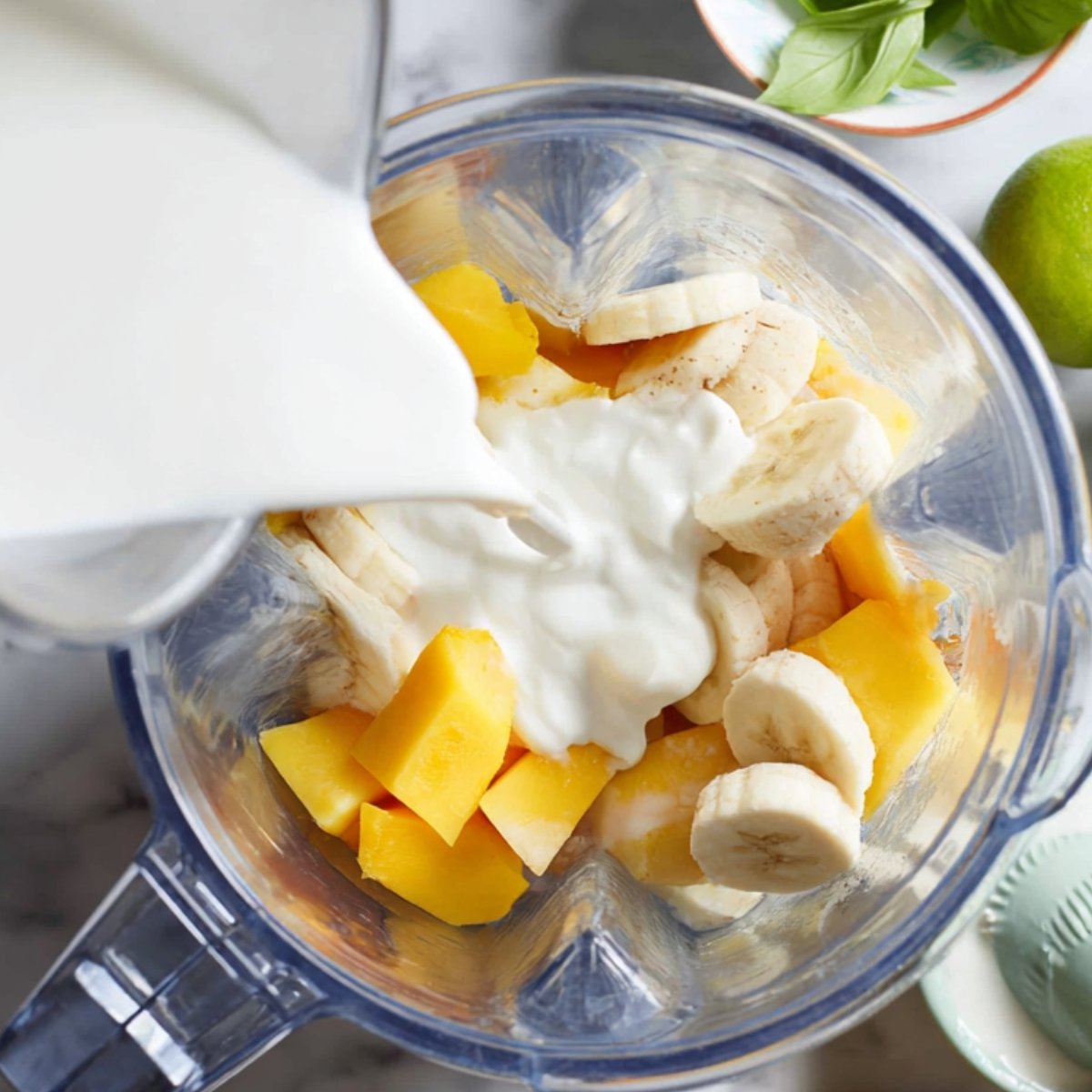 Overhead view of a blender jar filled with mango chunks and banana slices while yogurt is being poured in to make a smoothie.
