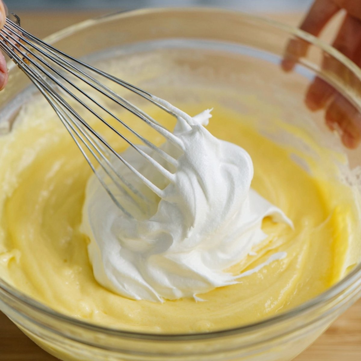 A whisk mixing whipped egg whites into a yellow pancake batter in a glass bowl.