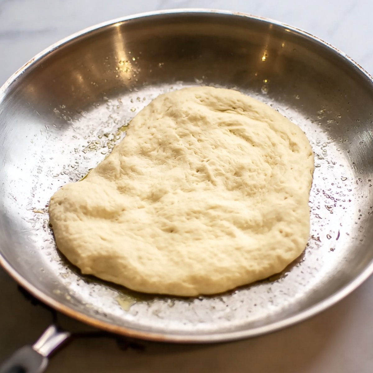 Close-up of naan dough cooking in a hot skillet. The dough has spread out into a rough, oval shape and is beginning to cook, with golden-brown spots forming along the edges.