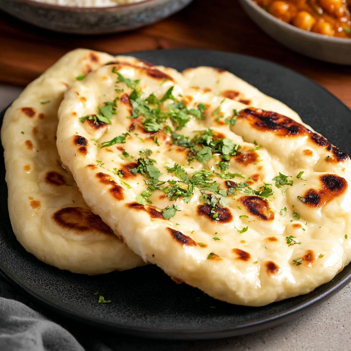 Two pieces of freshly cooked Homemade Naan Bread on a dark plate, topped with a sprinkle of fresh cilantro. The naan has a soft texture with golden-brown char marks and a slightly crispy edge.