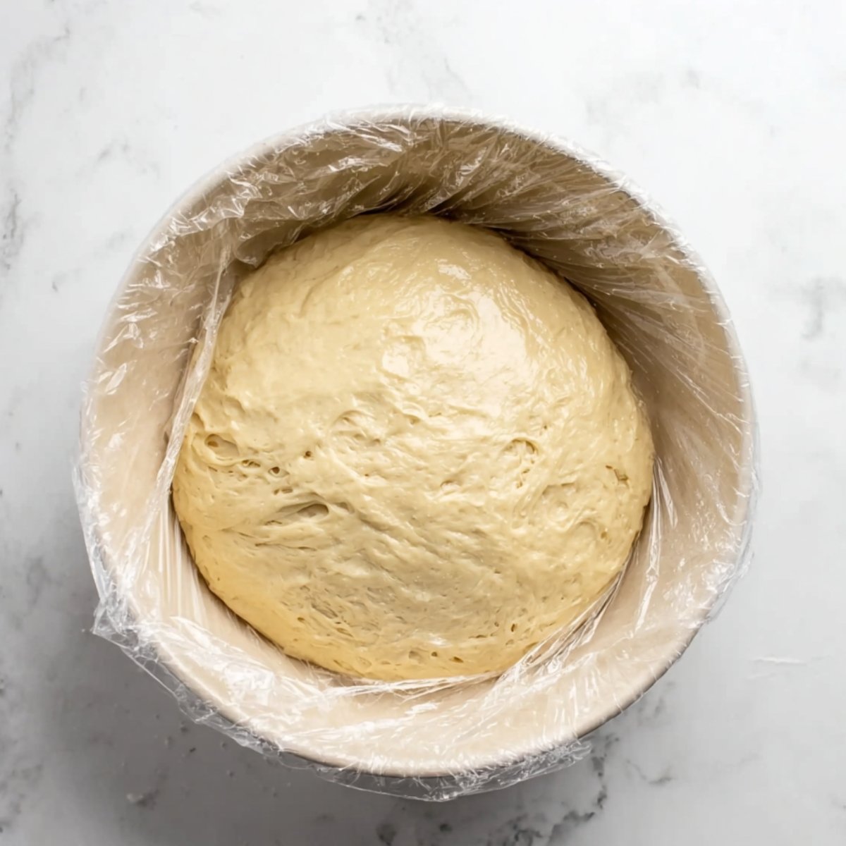 Overhead view of naan dough resting in a bowl, covered with plastic wrap. The dough has risen, creating a smooth, slightly shiny surface, indicating it's ready for the next step in the naan-making process.