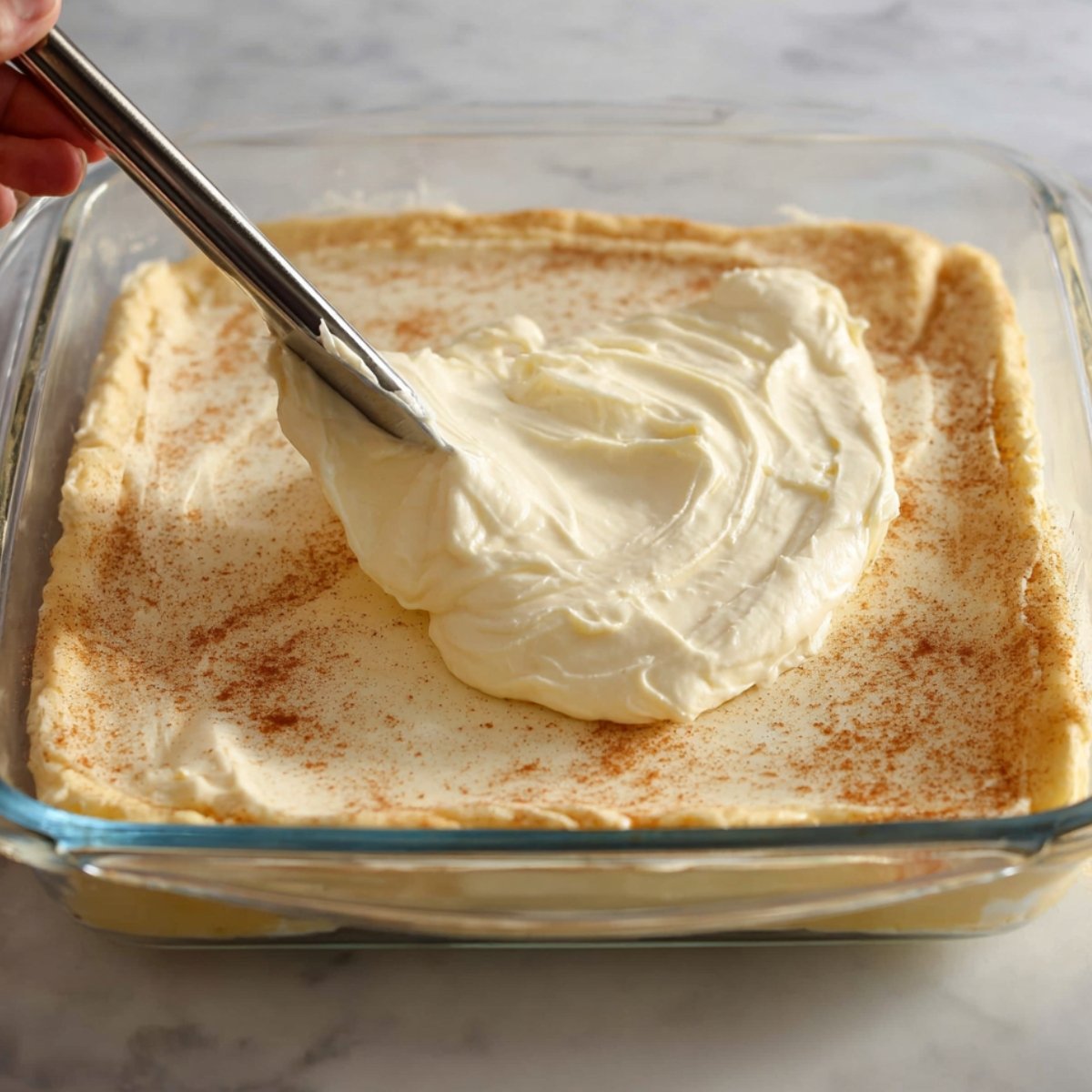 Spreading a creamy cheesecake filling over the dough in a glass baking dish, preparing it for the cinnamon-sugar topping.