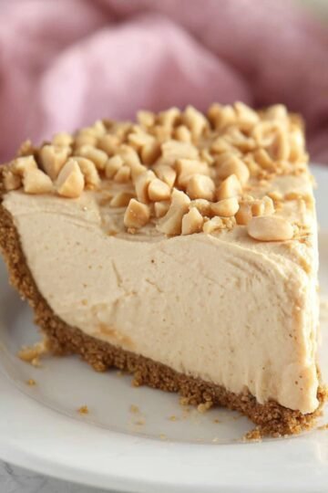 A slice of no-bake peanut butter pie on a white plate, topped with chopped peanuts and set against a soft pink background.