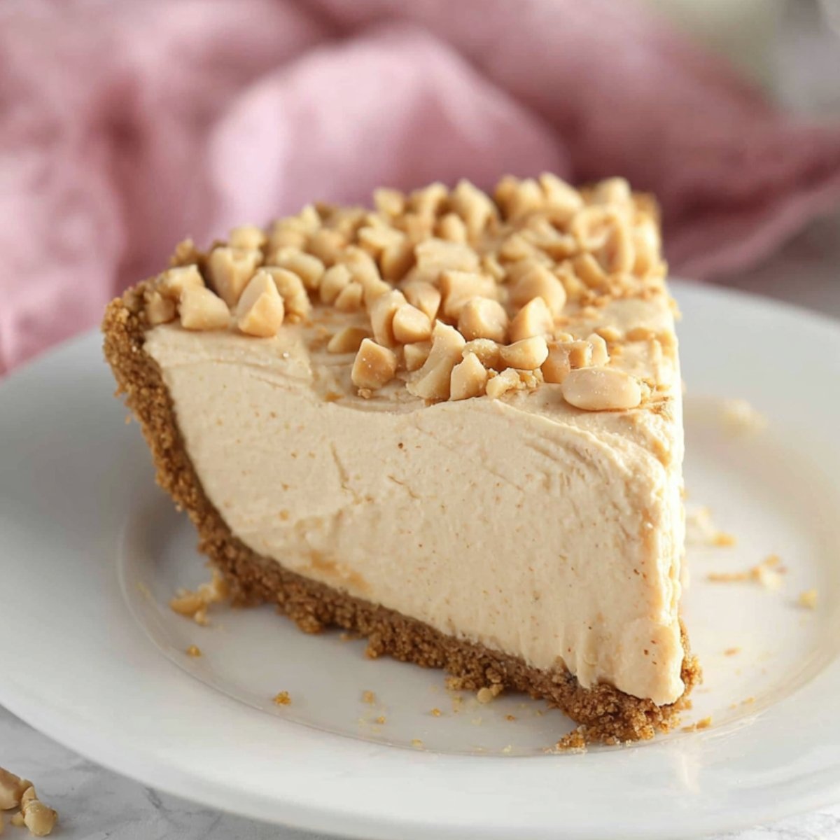 A slice of no-bake peanut butter pie on a white plate, topped with chopped peanuts and set against a soft pink background.