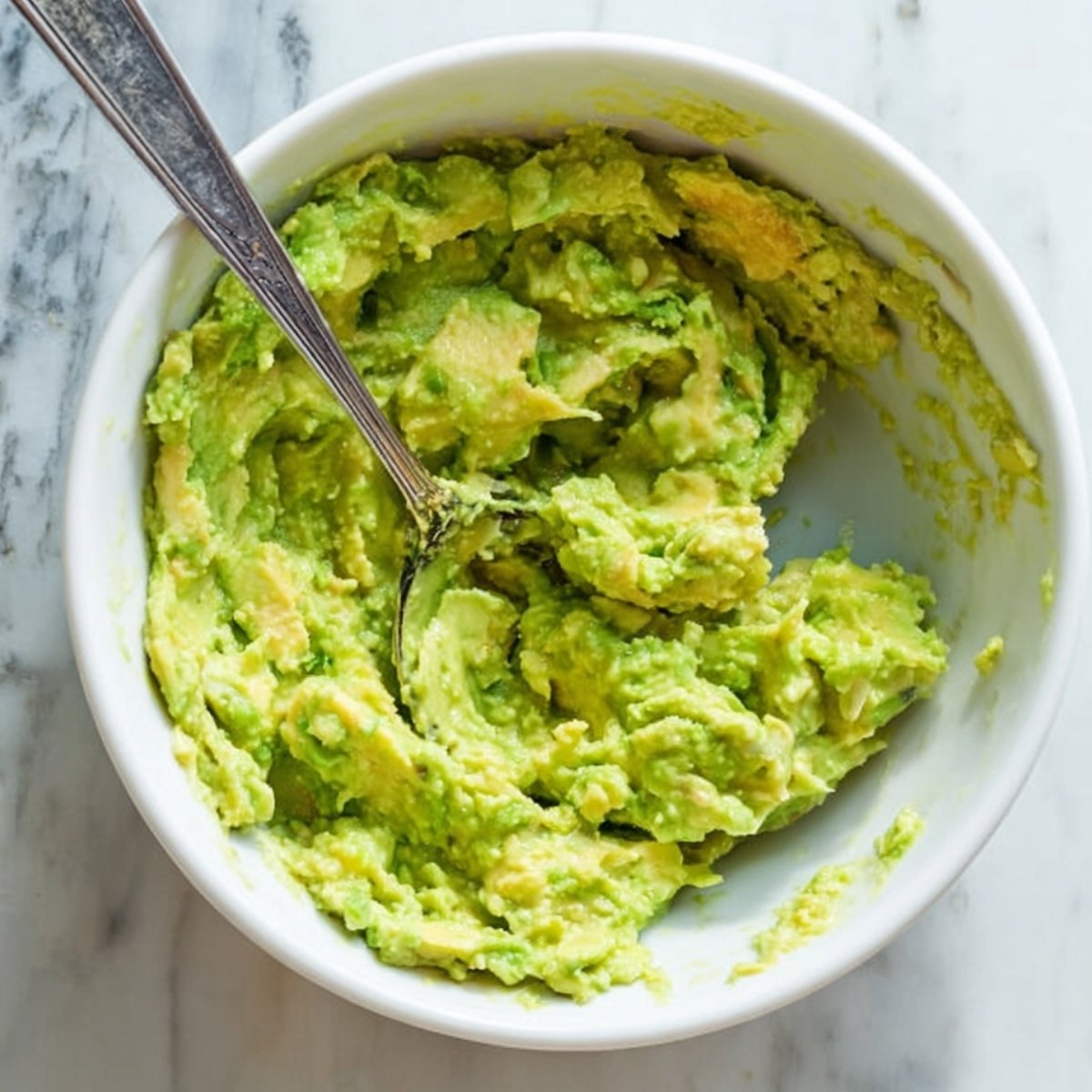 Bowl of freshly mashed avocado with a fork, showing chunky avocado texture on a marble countertop.
