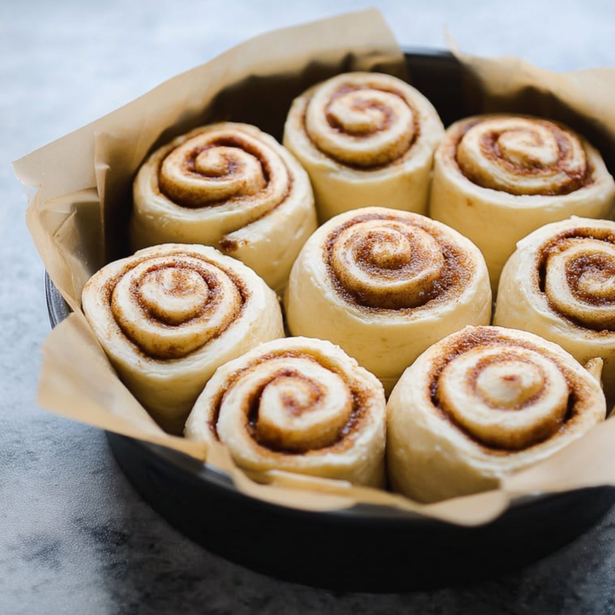 Freshly baked cinnamon rolls, swirled with cinnamon and topped with a generous layer of icing, resting on parchment paper in a pan.