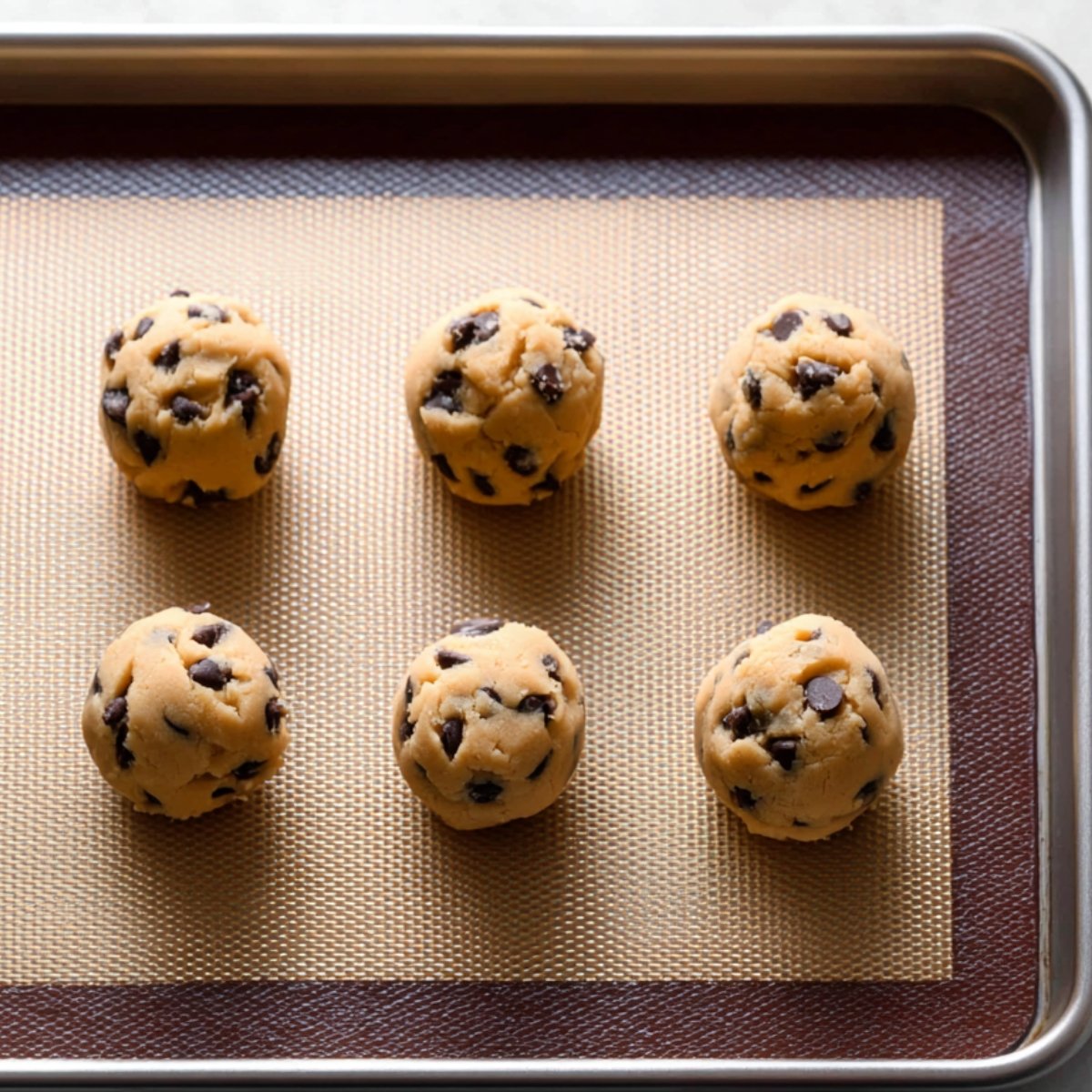 Chocolate chip cookie dough balls evenly spaced on a baking sheet lined with a silicone baking mat, ready to bake.