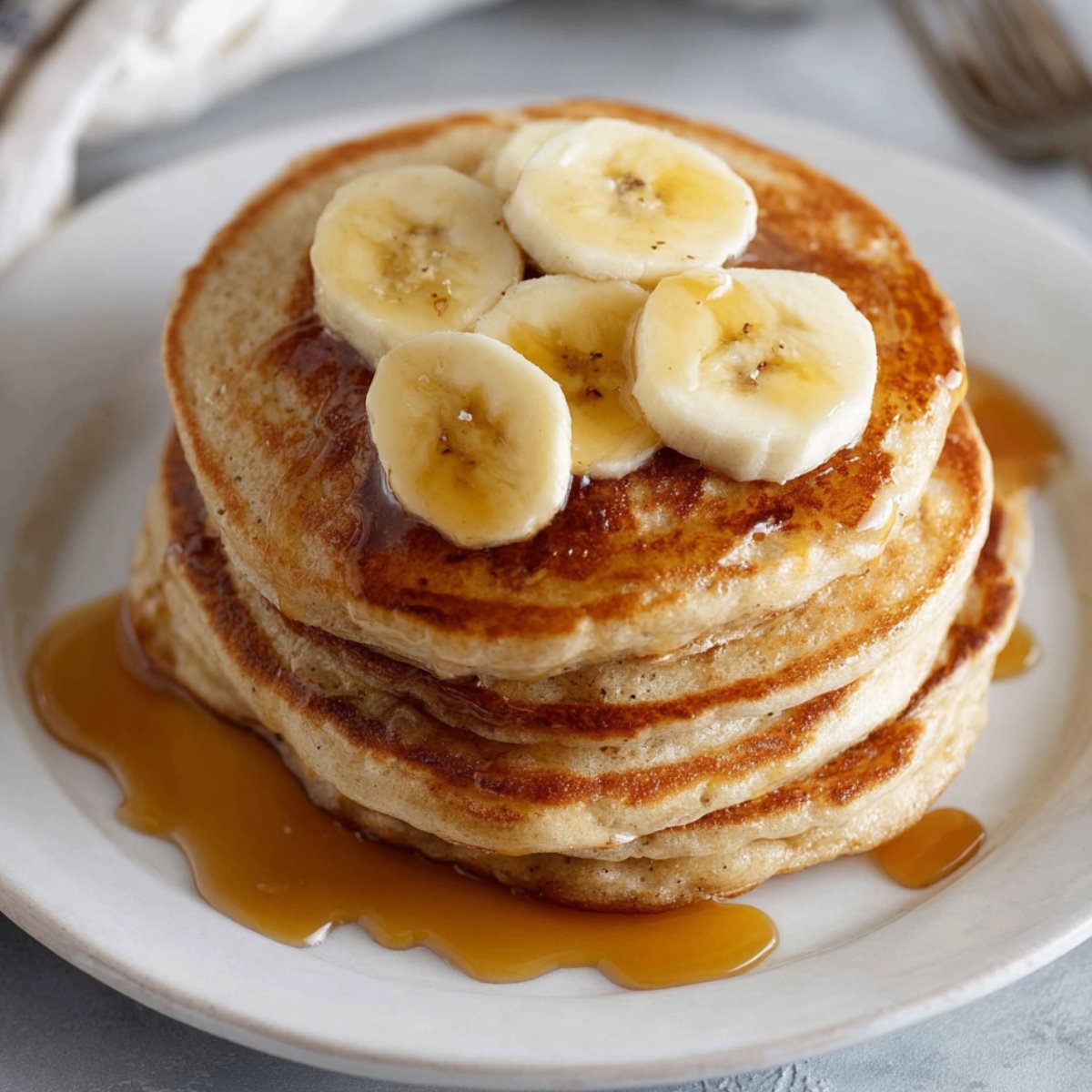 A stack of golden Fluffy Banana Pancakes topped with sliced banana and drizzled with syrup, served on a white plate.