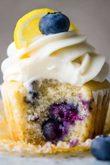 A frosted Lemon Blueberry Cupcakes with a bite taken out. The cupcake is topped with smooth frosting, a fresh blueberry, and a lemon slice, revealing a soft, moist interior with blueberries.