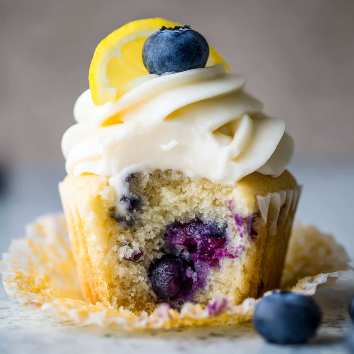 A frosted Lemon Blueberry Cupcakes with a bite taken out. The cupcake is topped with smooth frosting, a fresh blueberry, and a lemon slice, revealing a soft, moist interior with blueberries.
