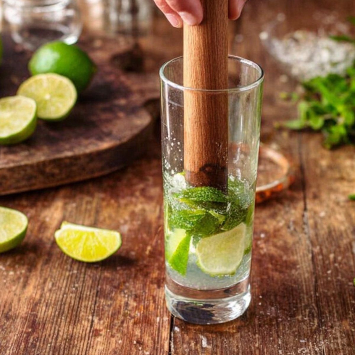 A wooden muddler is used to crush mint leaves and lime slices in a tall glass, preparing the base for a refreshing coconut mojito.
