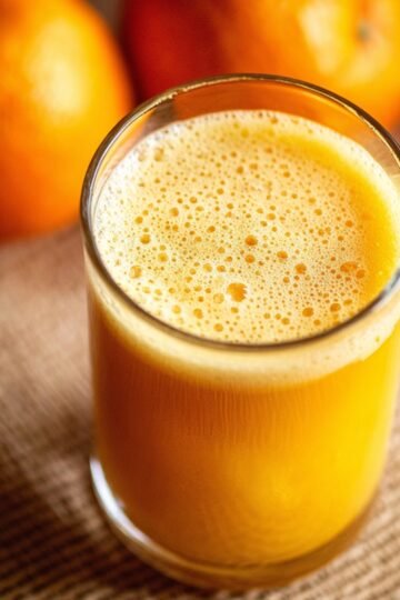 A glass of freshly squeezed orange juice with a foamy top, surrounded by whole oranges in the background.