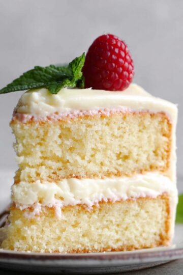 Slice of moist Vanilla Cake with creamy frosting, topped with a raspberry and mint leaf.